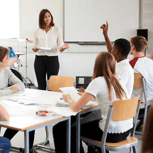 Blacbiblio | The ABC’s Toolkit is designed to help teachers create engaging lessons that are FUN and hands-on learning experiences. A teacher stands at the front of a classroom, engaging with six students seated at desks. One student raises their hand. Papers and supplies are on the desks, and a drum set is visible in the background. blacbiblio.com