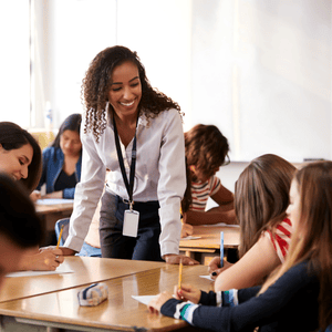 Blacbiblio | The ABC’s Toolkit is designed to help teachers create engaging lessons that are FUN and hands-on learning experiences. A teacher with curly hair and a lanyard leans over a desk, smiling while assisting students in a classroom. Students are engaged in writing tasks, seated at tables with supplies, in a brightly lit room. blacbiblio.com